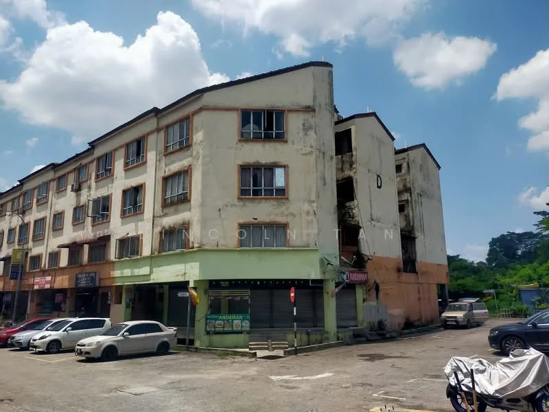 Exterior type: low-rise apartment block; features: weathered facade, cream painted facade, multiple storeys, ground floor commercial units, parking lot in front, visible signage for businesses, surrounding greenery, asphalt parking surface; style: traditional.
