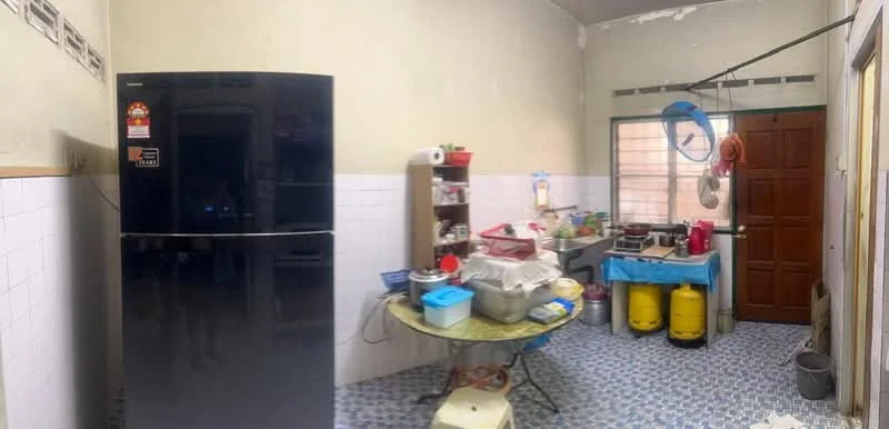 Kitchen features: black fridge, round dining table, blue tablecloth, gas stove, kitchen cabinets, white tiled walls, blue patterned floor tiles, wooden door, window with grid, various kitchen appliances, yellow gas cylinders, hanging utensils.