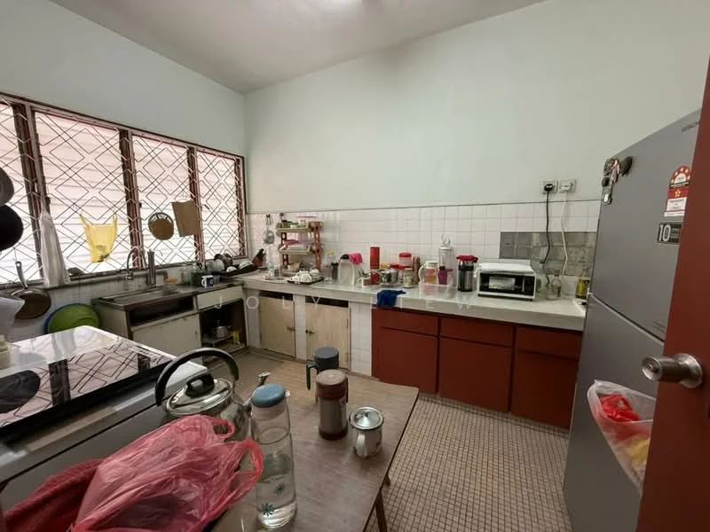 Kitchen features: white tiled walls, grey tiled backsplash, brown wooden cabinetry, stainless-steel sink, chrome faucet, built-in gas stove, freestanding refrigerator, microwave, toaster, kettle, countertop with clutter, window with metal grilles, floor tiles, open shelving, various kitchen utensils.
