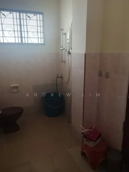 Bathroom features: white tiled walls, light beige tiled flooring, toilet, shower area with handheld shower, shower head, water heater, blue bucket, pink storage basket, small stool, window with decorative grille.