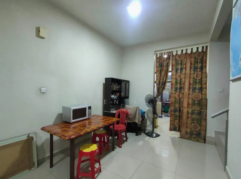 Room features: white painted walls, glossy tile flooring, dining table with red plastic chairs, microwave, curtains with floral pattern, standing fan, open space layout, staircase visible in background.