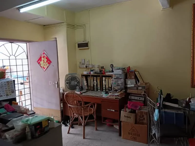 Room features: yellow painted walls, concrete flooring, wooden desk, office chair, bookshelves, file organizers, wall-mounted air conditioning unit, window with metal grille, decorative wall frame, cardboard boxes, fan, cluttered workspace.