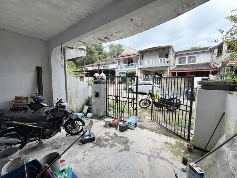 Exterior type: linked terrace house; features: concrete flooring, metal gate, surrounding fence, parking space for motorcycles, outdoor storage containers, visible neighboring houses, driveway, manicured greenery in background; style: traditional.