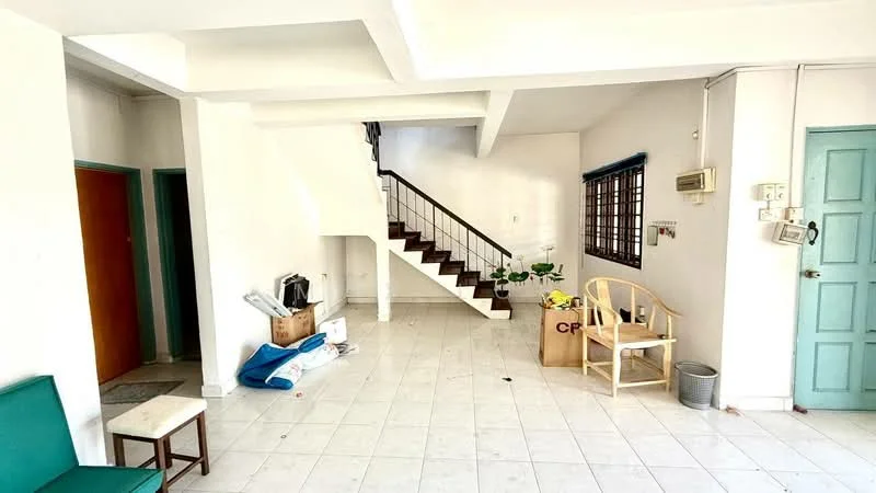 Room features: white painted walls, tile flooring, staircase with metal railing, large windows, doorway leading to another room, green door, wooden chair, small side table, cardboard boxes, indoor plants, air conditioning unit, wall-mounted light switch; style: minimalist.