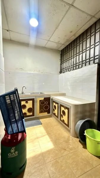 Kitchen features: single-bowl stainless-steel sink, chrome faucet, laminate countertop, white tiled backsplash, wood cabinetry with decorative panels, floor tiles in beige, overhead fluorescent lighting, gas cylinder, plastic waste bin, rubber mat.