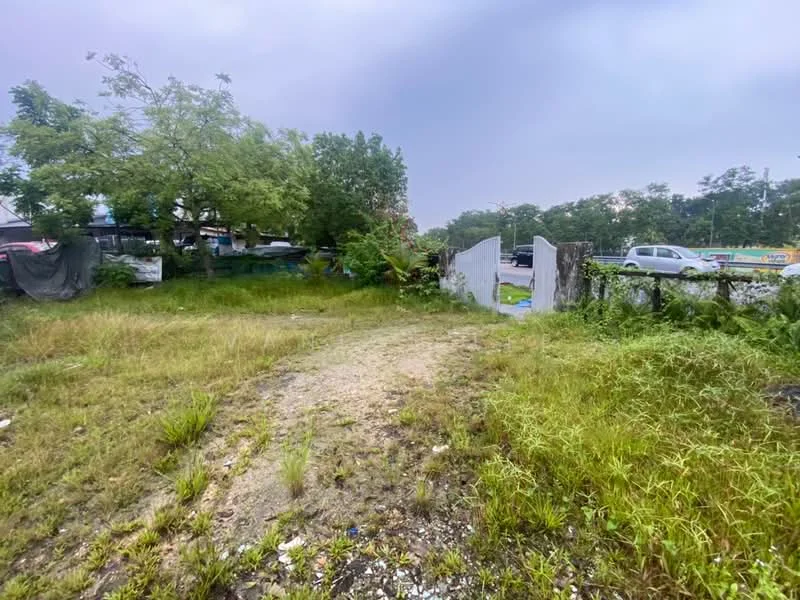 Exterior type: land; features: overgrown grass, dirt pathway, fenced perimeter, surrounding trees, adjacent road, urban context, visible vehicles, partially cleared area.