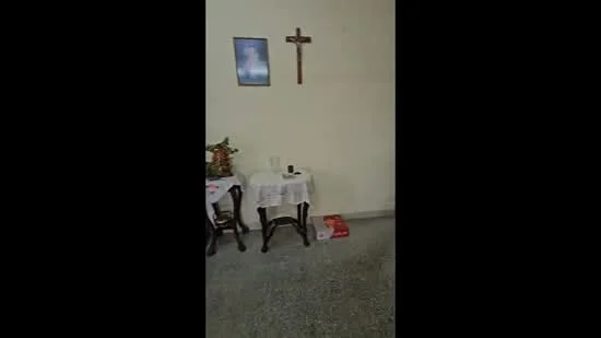 Room features: plain white walls, grey flooring, wooden side table, tablecloth on side table, wall-mounted crucifix, framed artwork on wall, decorative items on table; style: traditional.