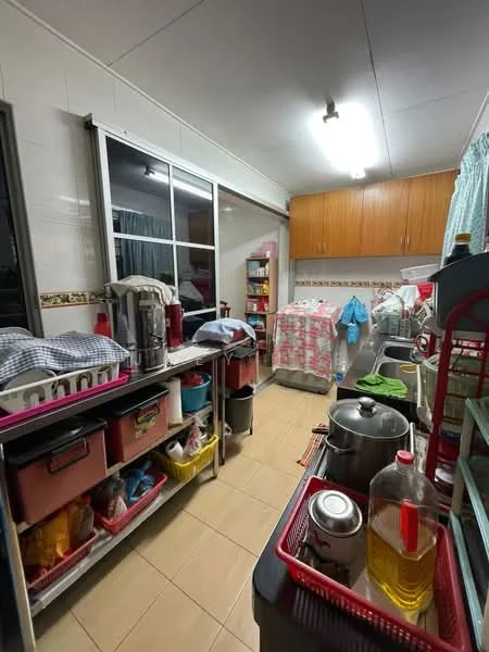 Kitchen features: white tiled walls, cream tiled flooring, wood cabinetry, stainless-steel sink, chrome faucet, kitchen countertop, multiple storage shelves, various kitchen appliances, open shelving, plastic storage bins, cooking pots, bottled cooking oil, window with sheer curtains, refrigerator, laundry area visible.