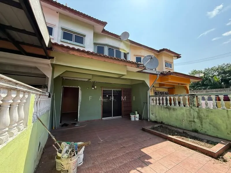 Exterior type: linked terrace house; features: two-storey building, green painted facade, terracotta roof tiles, balcony on upper floor, paved outdoor area, visible drainage system, satellite dishes on roof, manicured lawn area, fenced perimeter, outdoor storage area, windows with grills; style: modern.