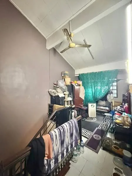 Room features: grey painted walls, sloped ceiling, ceiling fan, wooden staircase, decorative green curtain, area rug with geometric pattern, various storage items, clothing on railing, wooden furniture, plastic bottles, miscellaneous decor items.