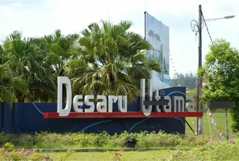 Exterior type: main entrance gate; features: signage with 'Desaru Utama', landscaping with palm trees, manicured lawn, decorative flower beds, blue painted wall, red accent at the base of the signage, street lighting, surrounding greenery; style: modern.
