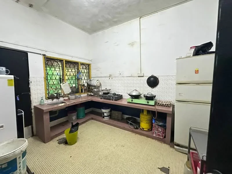 Kitchen features: white tiled walls, grey countertop, double-bowl stainless-steel sink, chrome pull-down faucet, freestanding refrigerator, freestanding stove, kitchen cabinets, window with security bars, vinyl flooring, multiple cooking pots, yellow bucket, green trash bin.
