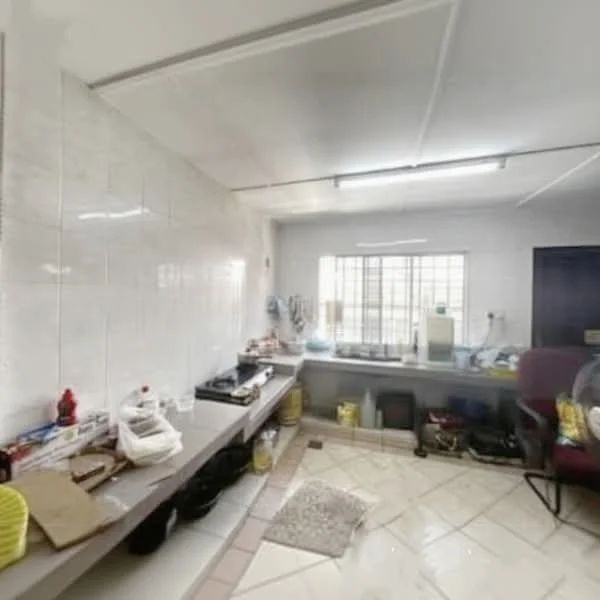 Room features: white tiled walls, light grey ceiling, fluorescent lighting, ceramic tile flooring, window with metal grilles, worktable with storage underneath, various kitchen appliances, shelves with kitchen items, plastic containers, office chair, sink, cleaning supplies; style: functional.