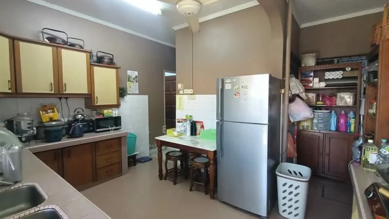 Kitchen features: brown wooden cabinetry, white tiled backsplash, double-bowl stainless-steel sink, chrome pull-down faucet, stainless-steel fridge, wooden dining table with two stools, open shelving with storage boxes, laundry basket, kitchen appliances including rice cooker and blender, cream painted walls, ceiling fan; style: traditional.