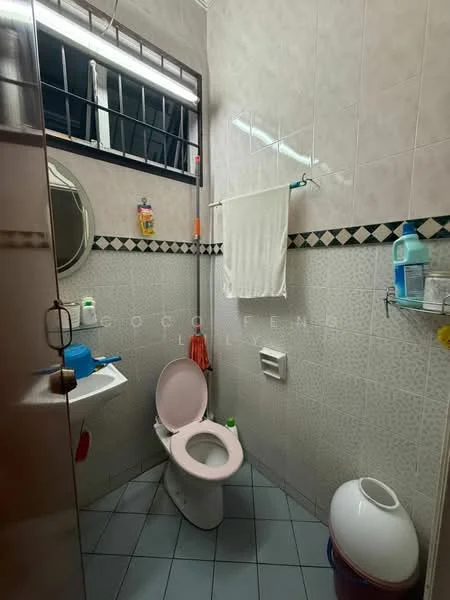Bathroom features: pink toilet, round mirror, white towel hanging on rack, blue sink, white soap dispenser, green bottle on sink, grey tiled flooring, white waste bin, shower area with glass partition, wall-mounted shelf with toiletries, decorative tile border.