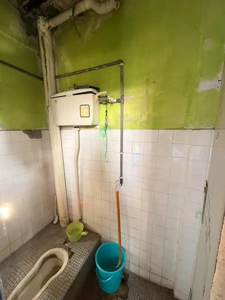 Bathroom features: green painted walls, white tiled walls, mosaic tile flooring, squat toilet, wall-mounted water tank, exposed plumbing, bucket, drainage pipe.