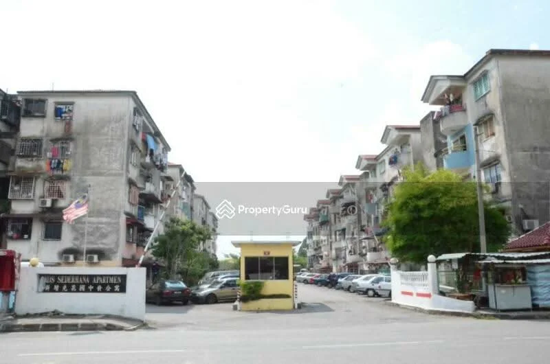 Exterior type: low-rise apartment block; features: multiple storeys, balconies with railings, exposed concrete facade, parking area, entrance gate, landscaped greenery, signage at entrance, surrounding residential area; style: traditional.