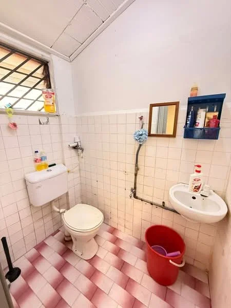 Bathroom features: white tiled walls, pink and white checkered tile flooring, one-piece toilet, sink with wall-mounted faucet, mirror above sink, wall-mounted storage shelf, window with metal grille, cleaning supplies on shelf, red bucket, soap dispenser, liquid soap bottle.