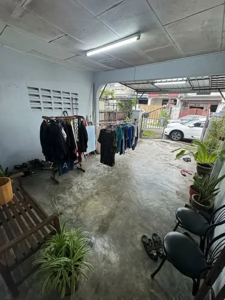 Room features: concrete flooring, white painted walls, fluorescent ceiling light, clothes rack with hanging clothes, potted plants, wooden bench, two black chairs, open space layout, metal gate, visible outdoor area.
