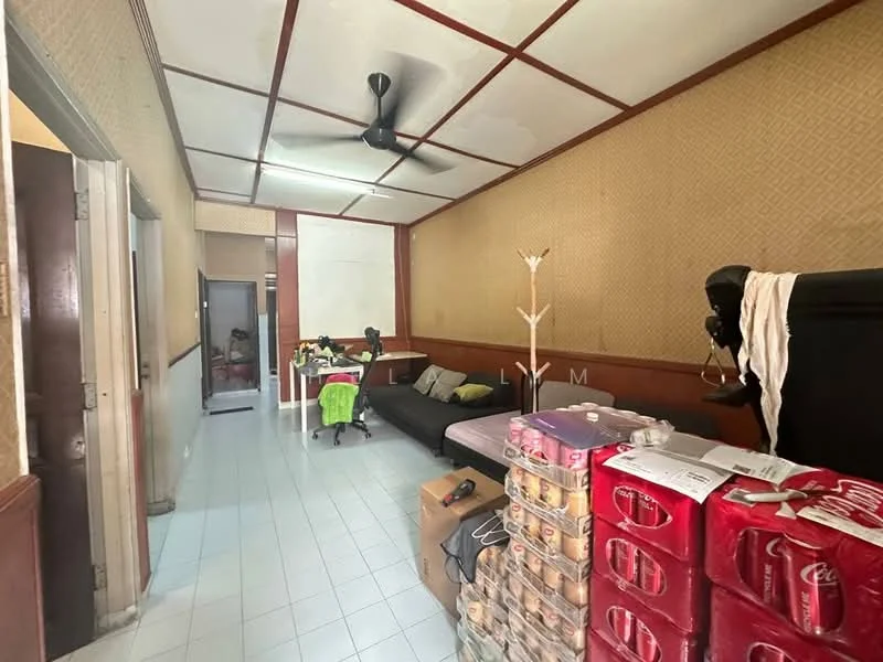 Living features: white tiled flooring, cream wallpaper with geometric pattern, wood paneling on walls, ceiling with wooden beams, 5-blade ceiling fan, large sectional sofa in dark color, coffee table, decorative floor lamp, open shelving unit, boxes of beverages stacked, table with chairs in background; style: traditional. Room features: white tiled flooring, cream wallpaper, wood paneling, doorway leading to another room.