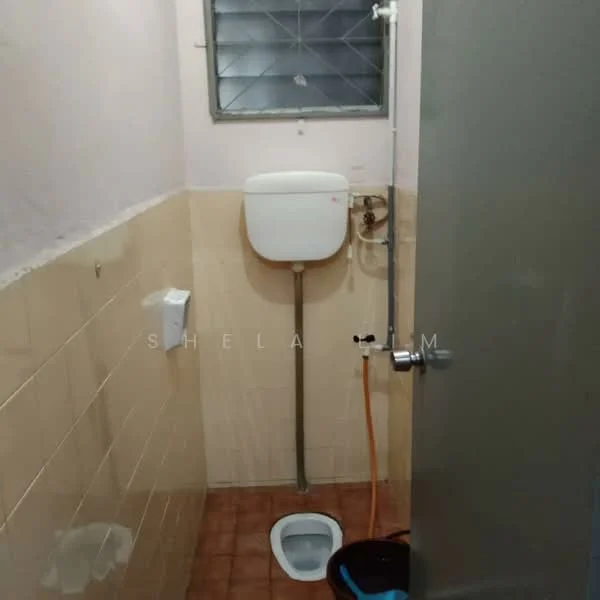 Bathroom features: white toilet, wall-mounted cistern, ceramic floor tiles, beige wall tiles, window with metal grille, shower pipe, toilet paper holder, cleaning bucket.