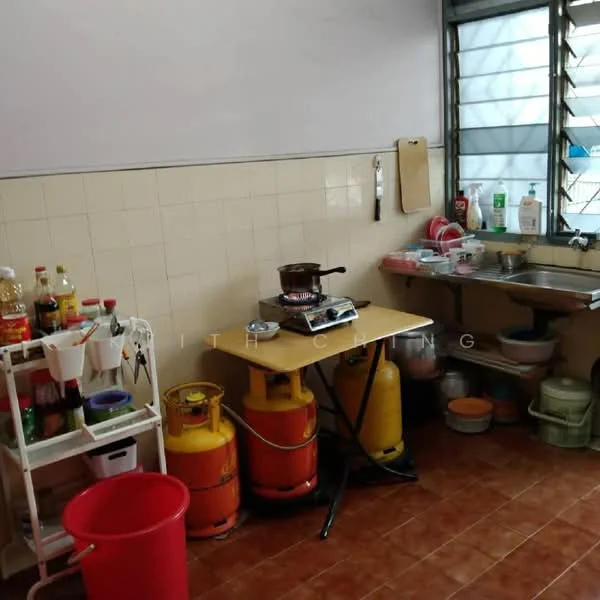 Kitchen features: tile flooring in terracotta color, white tiled walls, stainless-steel sink, single-bowl sink, wooden countertop, gas stove with pot, two gas cylinders, plastic storage containers, cleaning supplies on countertop, foldable table, red plastic trash bin, window with metal grilles.