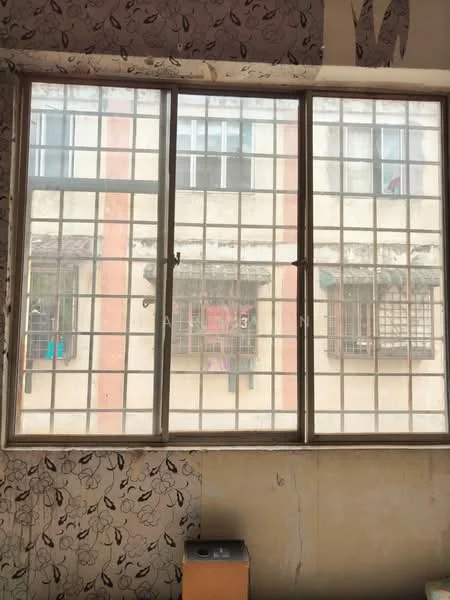Room features: large window with grid pattern, decorative wallpaper with floral pattern, bare wall with unfinished surface, wooden baseboard, visible external building with windows, metal grilles on external windows.
