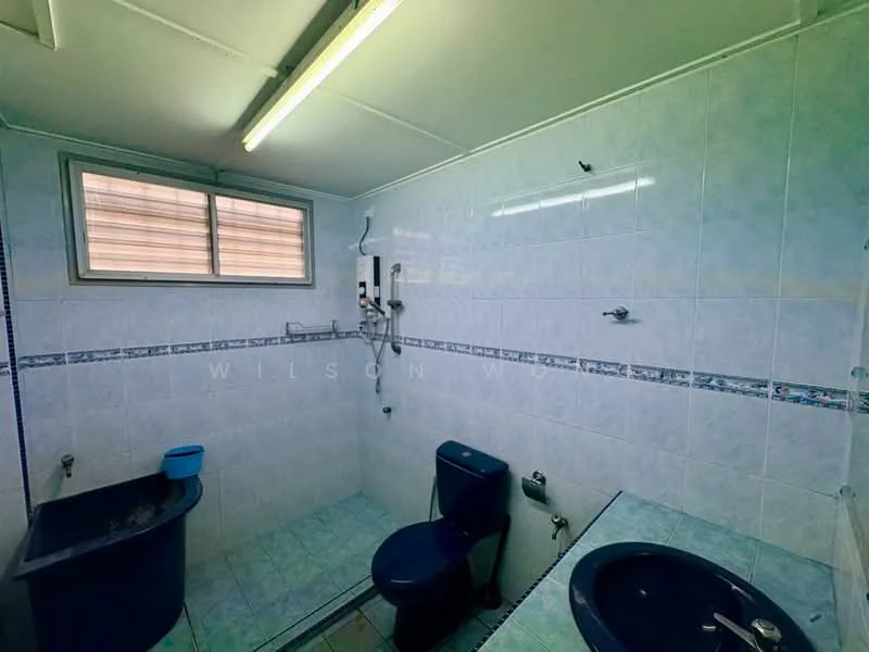 Bathroom features: white tiled walls, light blue tiled flooring, single-sink vanity with blue basin, one-piece toilet in blue, shower with wall-mounted showerhead, window for natural light, overhead fluorescent lighting, blue plastic bucket.