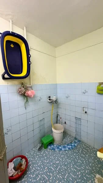 Bathroom features: blue tiled walls, light blue tiled flooring, toilet, sink with faucet, shower head, hanging storage basket, plastic bucket, laundry basket, yellow plastic container, exposed plumbing, wall-mounted soap holder.