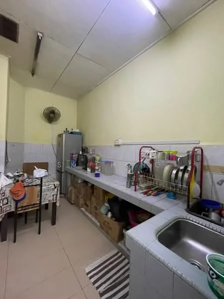 Kitchen features: yellow painted walls, tile backsplash, grey countertop, stainless-steel sink, chrome faucet, wall-mounted fan, refrigerator, kitchen cabinets, open shelving, dining table with tablecloth, various kitchen utensils, storage boxes, striped floor mat.