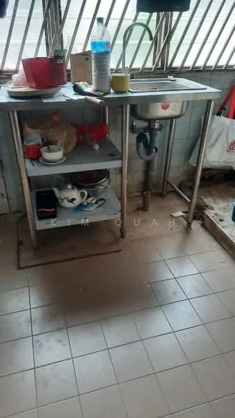 Kitchen features: stainless-steel sink, gooseneck faucet, metal shelving unit, red plastic bowl, white ceramic plates, glass bottle, yellow mug, dirty floor tiles, window with metal bars, cluttered countertop.