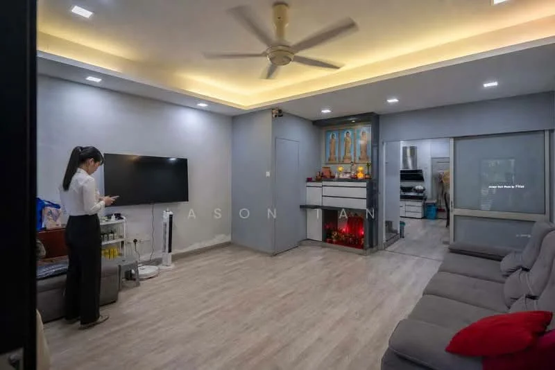 Living features: light grey painted walls, wooden flooring, tray ceiling with recessed lighting, 5-blade ceiling fan, wall-mounted TV, sofa with multiple seats in grey, coffee table, decorative wall with religious icons, open kitchen area visible, sliding glass door leading to outdoor space, LED lighting under cabinetry; style: modern.