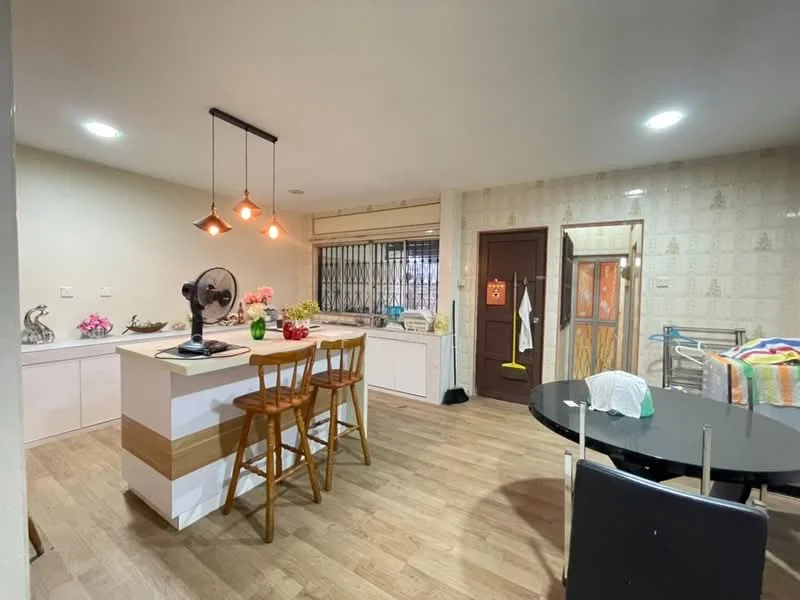 Kitchen features: wooden flooring, white cabinetry, kitchen island with breakfast bar, 3 wooden bar stools, pendant lighting, stainless-steel sink, chrome faucet, open shelving, decorative plants, kitchen appliances visible, window with security bars, wall-mounted fan; style: modern. Dining features: round dining table, black dining chairs, colorful tablecloth, decorative items on table; style: modern.