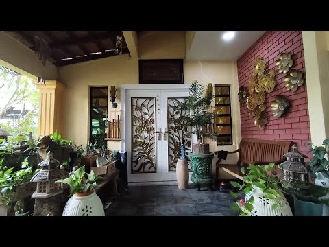 Exterior type: detached bungalow; features: double entrance doors with decorative design, brick accent wall in red, stone flooring, wooden ceiling with exposed beams, potted plants in various sizes, decorative wall art, bench seating, outdoor lighting, window treatments with decorative elements; style: traditional.