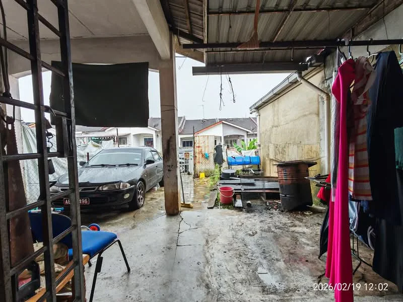 Exterior features: concrete flooring, metal roof, wooden support beams, clothes hanging on a line, blue plastic chair, parked car, outdoor storage area, visible neighboring houses, garden area with plants, water container, open space, partially covered area.