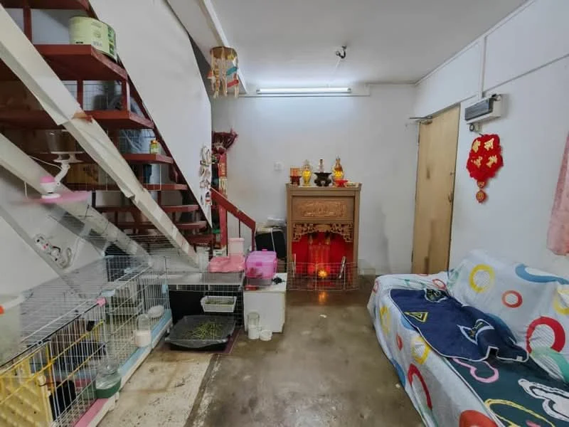 Living features: concrete flooring, white painted walls, staircase with wooden steps, sofa with colorful circular pattern, decorative altar with offerings, wall-mounted air conditioning unit, shelves for storage, cages for pets, small table, curtains with floral pattern.