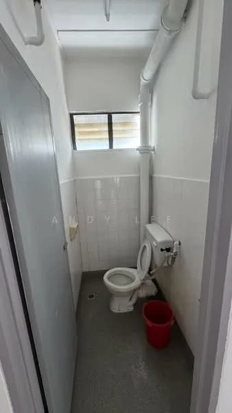 Bathroom features: white tiled walls, grey flooring, one-piece toilet, small window, plumbing pipes exposed, toilet paper holder, red bucket.