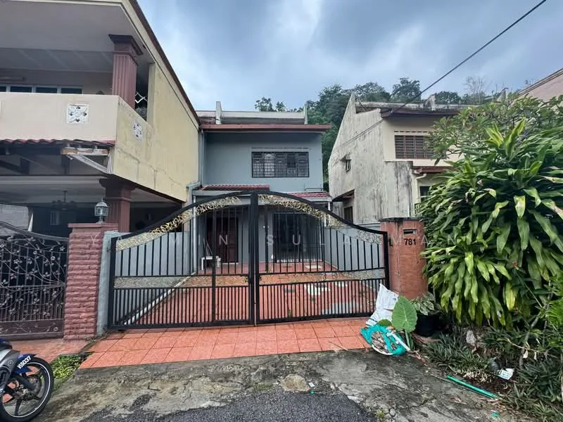Exterior type: semi-detached house; features: gated entrance with wrought iron gate, brick fence, tiled driveway, two-storey structure, exterior walls painted in light grey, red tiled roof, landscaped garden with shrubs, visible parking area, surrounded by greenery; style: modern.