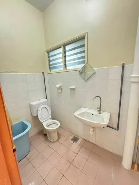 Bathroom features: white tiled walls, light beige tiled flooring, one-piece toilet, wall-mounted sink, chrome faucet, mirror, window with horizontal slats, toilet paper holder, soap holder, blue plastic bin.