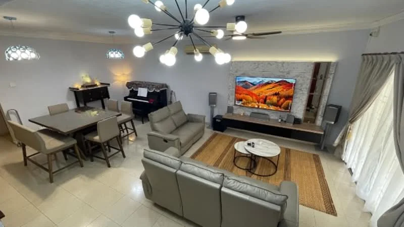 Living features: 3-seater fabric sofa in light grey, 2-seater fabric sofa in light grey, coffee table with round top, area rug with geometric pattern, wall-mounted TV, entertainment unit with storage, pendant light fixture with multiple bulbs, ceiling fan, large windows with sheer curtains, decorative wall with stone cladding, dining table with 6 chairs, piano; style: modern.