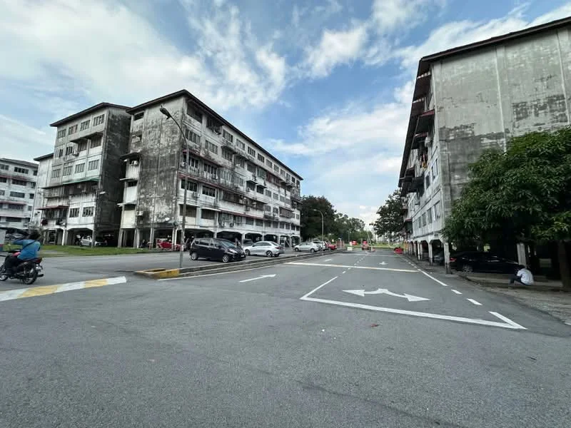Exterior type: low-rise apartment block; features: weathered concrete facade, multiple storeys, balconies with railings, parking spaces visible, surrounding greenery, street view, road with lane markings, sidewalk, nearby trees; style: traditional.