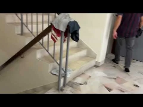 Room features: marble flooring, staircase with metal railing, neutral wall color, clothing items on stairs, doorway visible, natural light from nearby window; style: modern.