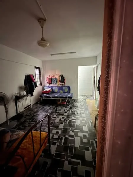 Room features: black and white patterned flooring, white painted walls, ceiling fan, single bed with yellow bedding, blue sofa, clothes hanging on wall-mounted hooks, two standing fans, doorway leading to bathroom, window with grid pattern, wooden door frame.