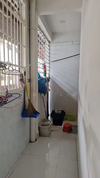 Room features: white painted walls, glossy tile flooring, window with security bars, cleaning supplies, broom, bucket, plastic container, laundry area, natural light from window.