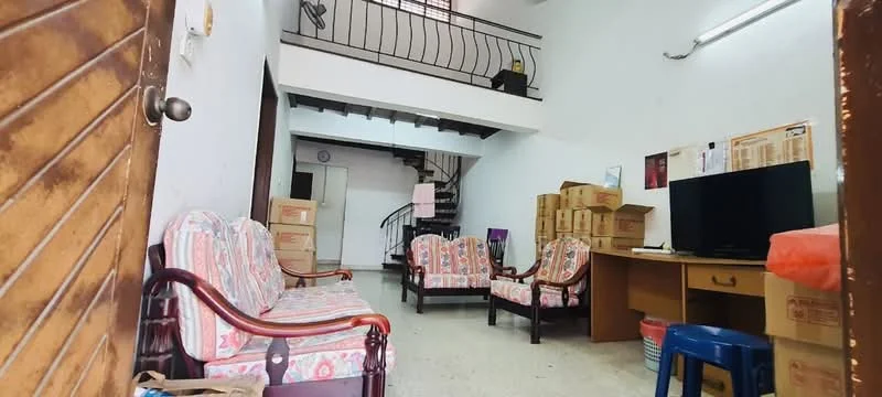 Living features: two 3-seater chairs with floral upholstery, wooden armchairs, coffee table, TV console, wall-mounted TV, staircase leading to upper floor, boxes stacked against the wall, white painted walls, tile flooring, overhead fluorescent lighting.