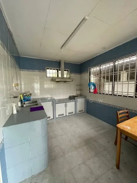 Kitchen features: blue tiled walls, white tiled backsplash, grey laminate countertop, stainless-steel sink, built-in cabinets, freestanding gas stove, exhaust hood, window with metal grille, table with wooden legs, plastic chair, grey tiled flooring, ceiling lights.