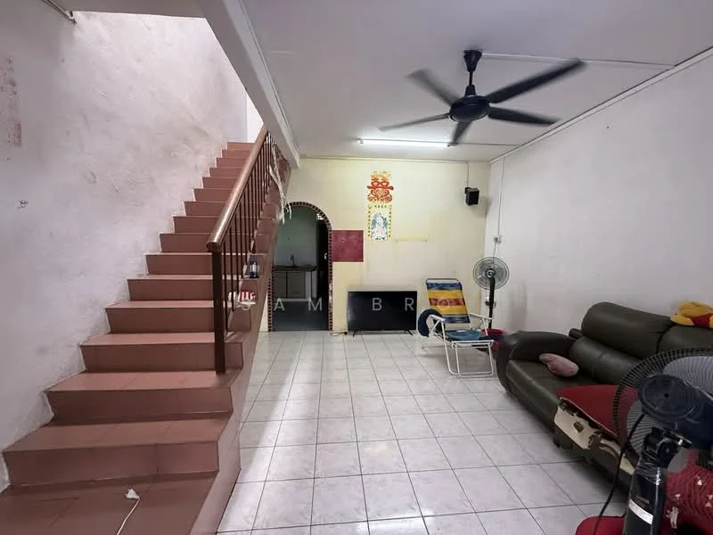 Living features: white painted walls, ceramic tile flooring, 5-blade ceiling fan, sofa in dark brown leather, coffee table, two standing fans, staircase with metal railing, area rug, decorative wall art. Room features: archway leading to another room, kitchen area visible in background.