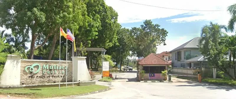 Exterior type: gated community; features: entrance gate with guard house, stone-clad entrance wall, flag poles with national flags, manicured landscaping, paved driveway, surrounding trees, residential buildings visible in background; style: modern.
