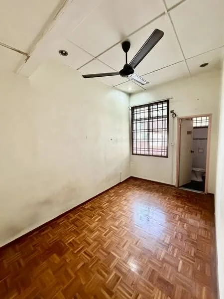 Room features: wooden flooring, white painted walls, ceiling fan, window with grille, door leading to bathroom, light fixture, corner wall, no visible furniture.