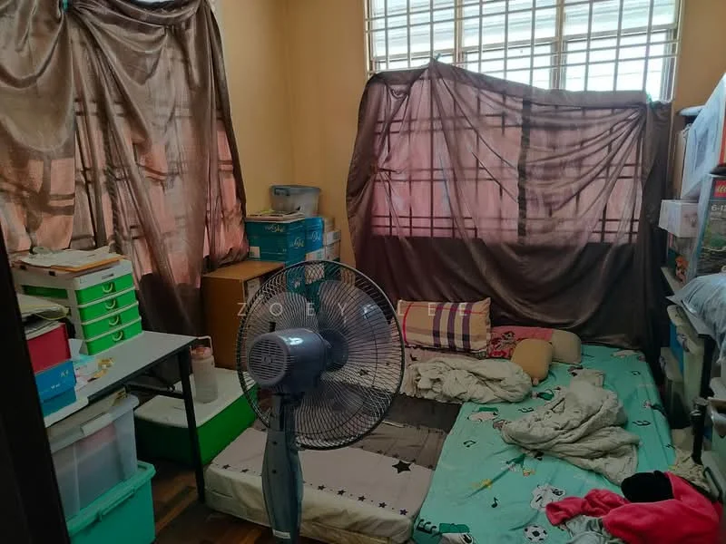 Room features: wooden flooring, cream painted walls, large window with grid pattern, floor fan, bed with bedding, scattered pillows, green storage drawers, boxes and clutter, curtains, unmade bed.